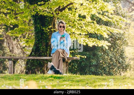 Femme portant des lunettes de soleil à l'aide d'un smartphone assis sur un banc près d'un chien de compagnie au parc Banque D'Images