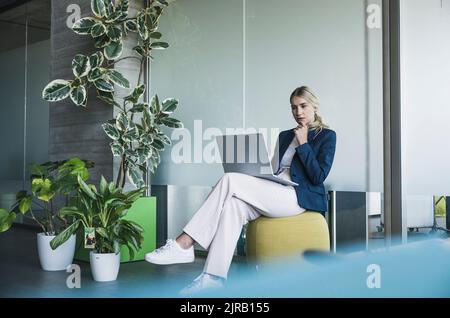 Femme d'affaires attentionnés avec ordinateur portable assise sur un tabouret de pouf dans le bureau Banque D'Images