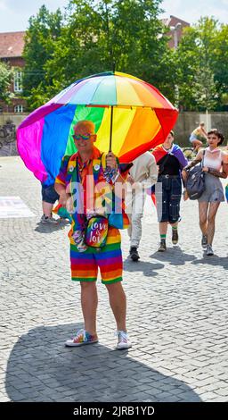 Braunschweig, Allemagne, 13 août 2022: Jeune homme blond avec lunettes de soleil, short et un parapluie avec cape dans les couleurs de l'arc-en-ciel sourit au b Banque D'Images