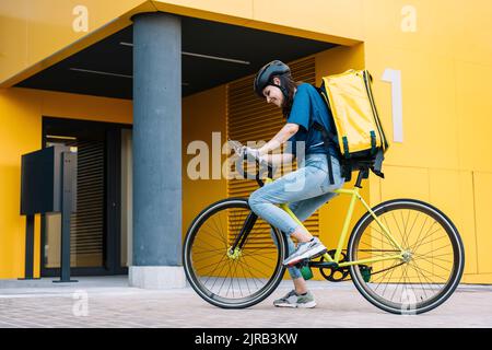 Bonne femme de livraison utilisant un smartphone assis sur un vélo devant le bâtiment Banque D'Images