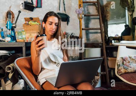 Un designer de mode tenant une tasse à café jetable à l'aide d'un ordinateur portable à l'atelier Banque D'Images