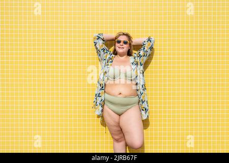 Femme souriante de taille plus avec les mains derrière la tête devant le mur jaune Banque D'Images