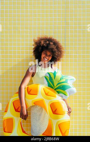 Fille souriante avec anneau gonflable debout devant le mur jaune Banque D'Images