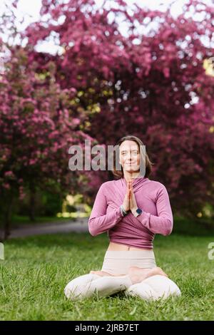 Femme souriante aux mains claspéties faisant du yoga assis dans un jardin de fleurs de pomme Banque D'Images