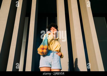 Femme avec les yeux fermés portant des oranges dans un sac en maille devant la colonne Banque D'Images