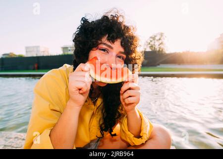 Jeune femme mangeant du melon d'eau par étang au coucher du soleil Banque D'Images