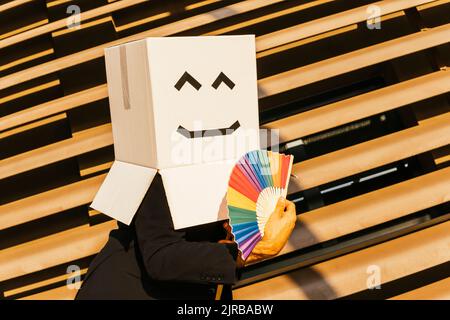 Homme d'affaires portant une boîte avec un visage souriant tenant un ventilateur de main de couleur arc-en-ciel le jour ensoleillé Banque D'Images
