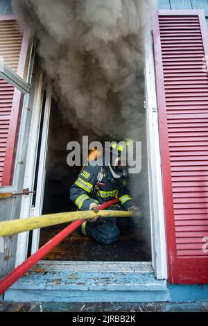 Un pompier du Service des incendies de Sag Harbor portant un appareil respiratoire s'agenouille dans la porte du bâtiment et alimente le tuyau à ses collègues pompiers f Banque D'Images