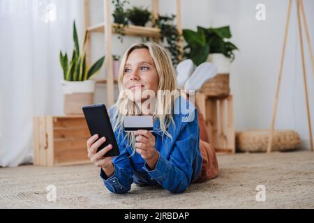 Femme contemplative faisant des achats en ligne sur le sol à la maison Banque D'Images