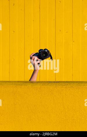 Main de la femme tenant l'appareil photo derrière le mur jaune Banque D'Images