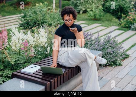 Jeune femme d'affaires envoyant la messagerie vocale par téléphone intelligent sur le banc au parc de bureau Banque D'Images