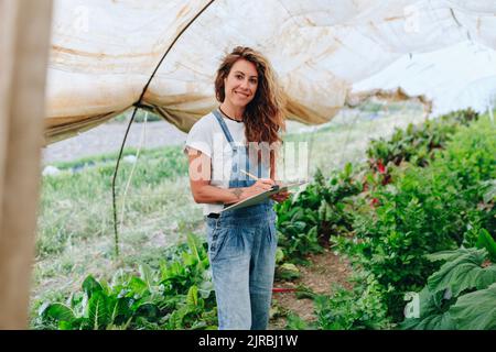 Un ouvrier agricole heureux avec un presse-papiers prenant l'inventaire des légumes à la serre Banque D'Images