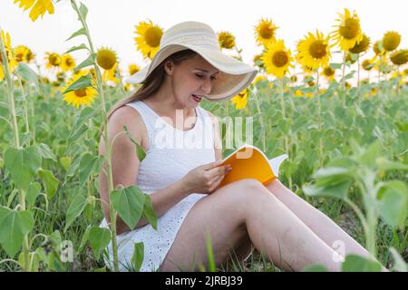 Femme avec chapeau lecture livre assis au champ de tournesol Banque D'Images