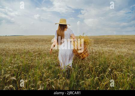 Une jeune femme dans un chapeau avec un panier en osier fait son chemin à travers un champ de blé. Vue arrière. Banque D'Images