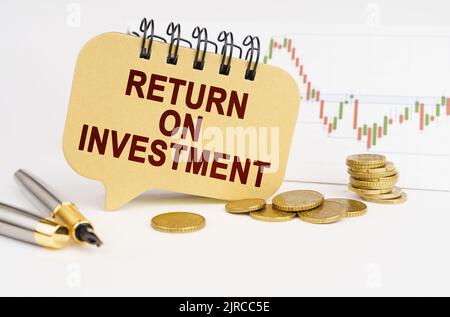 Concept d'entreprise. Sur la table se trouvent des pièces de monnaie, un stylo, un graphique et un signe avec l'inscription - RETOUR SUR INVESTISSEMENT Banque D'Images