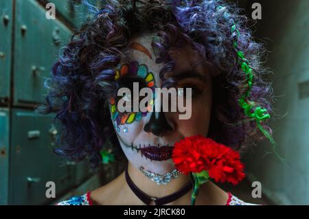 Portrait d'une jeune femme caucasienne avec la Calavera Catrina maquillage dans la crypte d'un cimetière, regardant la caméra en vue frontale, tenant un f rouge Banque D'Images