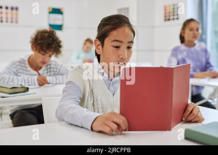 Portrait d'un adolescent assis à un bureau dans une salle de classe et livre de lecture avec un groupe d'enfants en arrière-plan Banque D'Images