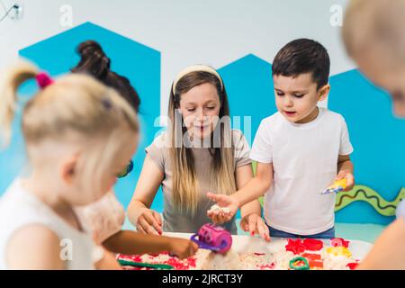 Détente sensorielle jeu avec sable cinétique moulable à l'école maternelle. Les tout-petits avec leur professeur s'amusent autour de la table en utilisant différents outils pour sculpter le sable, tels que des punaises texturées et colorées, des pinces coupantes, des étuis en silicone. Compétences motrices fines et développement créatif . Photo de haute qualité Banque D'Images