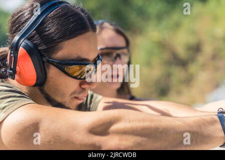Gros plan d'un homme de race blanche portant des lunettes de sécurité et des écouteurs visant le pistolet observé par un instructeur féminin. Prise de vue horizontale en extérieur. Photo de haute qualité Banque D'Images