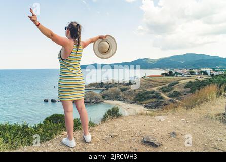 Jeune femme caucasienne joyeuse dans une robe rayée qui lui étire les bras tout en admirant le paysage marin grec magnifique. Concept de vacances. Photo de haute qualité Banque D'Images