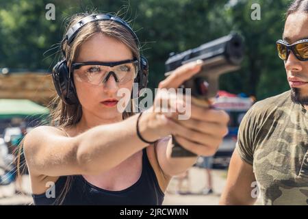 Jeune femme caucasienne portant des lunettes de protection et des écouteurs visant le pistolet observé par un instructeur barbu. Formation sur les armes à feu. Tir Horiozntal. Photo de haute qualité Banque D'Images
