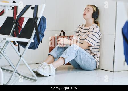 Portrait complet d'une adolescente dépressive assise sur le sol à l'école avec un casque Banque D'Images