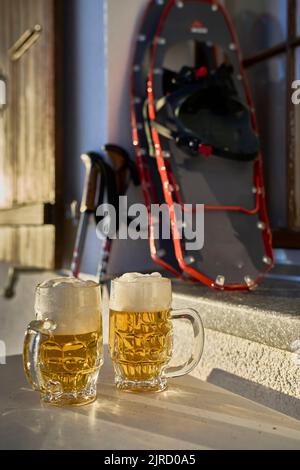 Deux grandes tasses de bière garrées se trouvent dans le soleil du soir sur la table, devant Mountain Hut. En arrière-plan des raquettes et des bâtons de ski flous. Riederalp, Valais, Banque D'Images