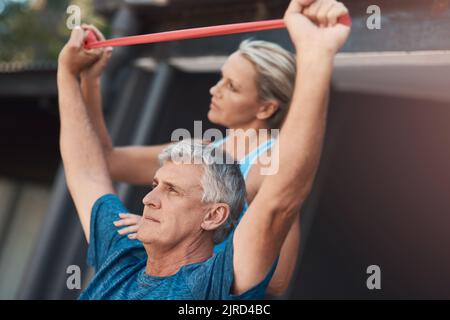 Restez motivé, peu importe. Un homme mûr étirant ses muscles de bras avec une bande élastique tout en étant aidé par une femme. Banque D'Images