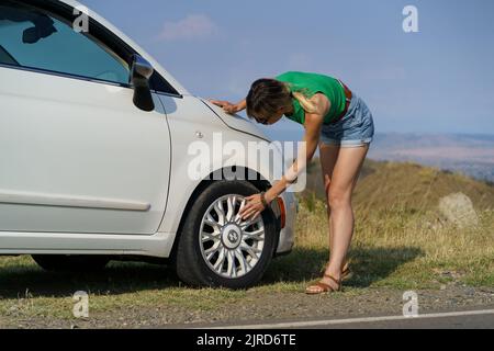 Une femme se penche en posant la main sur un pneu de roue pour vérifier l'état de la voiture blanche garée sur la route Banque D'Images