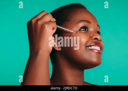 Portrait d'une jeune femme afro-américaine souriante appliquant du sérum facial avec compte-gouttes sur le visage, isolée sur fond turquoise. Sérum, huile pour le visage Banque D'Images