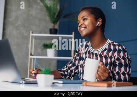 Jeune femme africaine américaine entrepreneur ayant un appel, assis à son bureau et de boire du café. Un esprit d'entreprise réussi. Appel professionnel. Banque D'Images