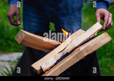 L'homme jette du bois de chauffage dans un brazier. Démarrer un feu de camp dans une fosse avec allumettes de sécurité et d'allumage. Camping Lifestyle.Spending temps libre concept, été Banque D'Images