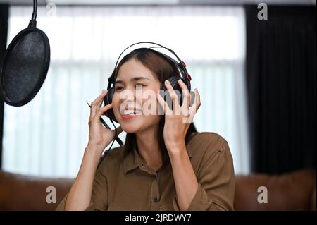 Jeune chanteuse asiatique attrayante et talentueuse, portant un casque, utilisant un microphone, chantant sa chanson de démonstration et enregistrant sa voix en studio. Banque D'Images