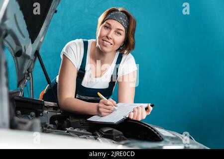 Le portrait d'une jeune femme mécanicien souriante dans une combinaison affiche des documents inclinant à la voiture. Le concept de l'égalité de la femme et travaille à l'auto réparation sho Banque D'Images