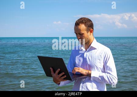 un homme avec un ordinateur portable dans la mer dans une chemise blanche de bureau. travail en vacances. Banque D'Images