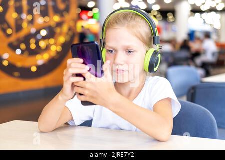 Une petite fille avec un casque regarde des dessins animés sur son téléphone assis à une table dans un café. Adorable écolière écoutant de la musique avec un téléphone portable et Banque D'Images