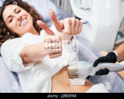 Jeune patient satisfait avec l'expression joyeuse du visage montrant les pouces vers le haut pendant SMAS - session de levage dans la clinique de médecine esthétique. Méthode non-opératoire rajeunissement de la peau du corps. Concentrez-vous sur les mains des femmes. Banque D'Images