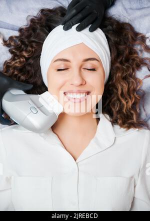 Vue de dessus de belle jeune femme avec les yeux fermés portant un bandeau blanc tout en recevant un soin du visage au centre de bien-être. Patiente gaie ayant une procédure de soulèvement du visage et souriant. Banque D'Images