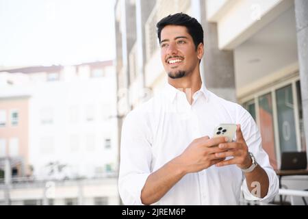 Joyeux et souriant latin sombre-cheveux homme dans la chemise blanche en utilisant le smartphone à l'extérieur, la messagerie texte, chat en ligne, la navigation sur le Web, beau homme avec le téléphone marchant dans la rue de la ville Banque D'Images