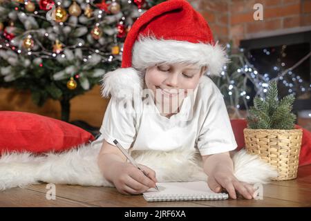Joyeux Noël aux enfants. Garçon enfant dans le chapeau du Père Noël écrit une lettre au Père Noël sous l'arbre de Noël avec des lumières de Noël, près de la cheminée. Famille Banque D'Images