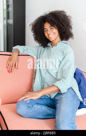 Portrait d'une femme attirante avec des cheveux bouclés assis sur le canapé dans un appartement moderne, une femme africaine multiraciale se reposant à la maison, regardant l'appareil photo et souriant avec un grand sourire Banque D'Images