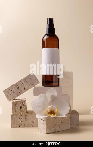 Bouteille cosmétique en verre foncé avec étiquette vierge près des fleurs d'orchidées blanches et des pierres sur jaune clair, gros plan, maquette. Produit de beauté fait main pour le soin de la peau, Banque D'Images