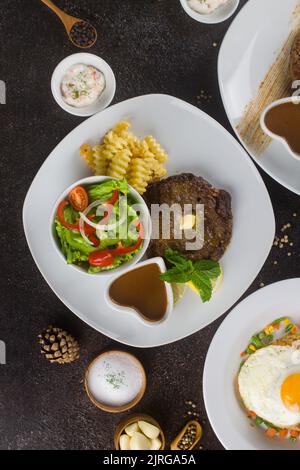 Steaks de bœuf grillés à la sauce aux pommes de terre, aux légumes et au steak sur une assiette blanche sur une table rustique texturée Banque D'Images