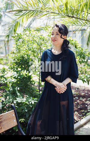 Belle japonaise heureuse d'âge moyen dans noir décontracté robe parc d'été, souriant. Lumière du soleil, espace de copie Banque D'Images