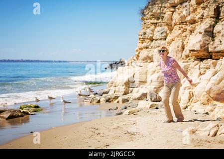 Une femme âgée en lunettes de soleil nourrit des mouettes sur la plage Banque D'Images