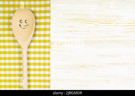 Cuillère en bois avec un sourire sur une nappe à carreaux jaunes sur une surface en bois blanc. Maquette pour menu ou recette, restaurant, site Internet avec cuisine. Cuisine f Banque D'Images