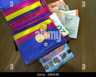 Un portefeuille en cuir coloré s'ouvre sur une table en bois. Quelques billets regardent et il y a trois pièces en euros sur le cuir. Banque D'Images