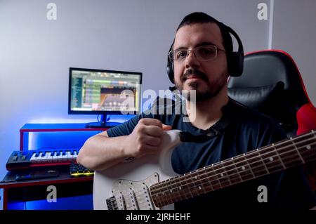 portrait du jeune homme du caucase du sud avec des lunettes et la barbe est assis et tenant sa guitare électrique dans son studio à la maison, regardant la caméra, l'esprit Banque D'Images