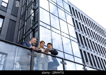 Trois hommes d'affaires femme et homme, à l'extérieur du bâtiment de bureau, sérieux et réfléchi discuter et regarder la tablette électronique, homme asiatique et femme dans les costumes d'affaires Banque D'Images