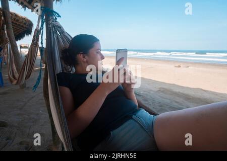 Belle jeune femme hispanique qui parle sur son téléphone portable, vêtue d'un t-shirt bleu reposant dans un hamac près de la plage par temps ensoleillé Banque D'Images